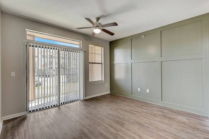 Bright apartment room featuring elegant wood flooring, stylish green accent wall, and large sliding glass doors with vertical blinds for abundant natural light.