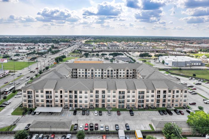 Elegant aerial view of a modern apartment complex with spacious parking, set against a vibrant cityscape and a beautiful sky backdrop.