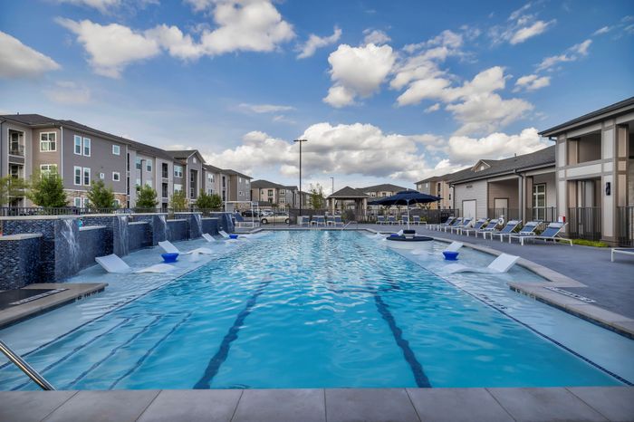 Luxurious apartment complex with inviting swimming pool, chic lounge chairs, and modern architecture under a bright, blue sky.