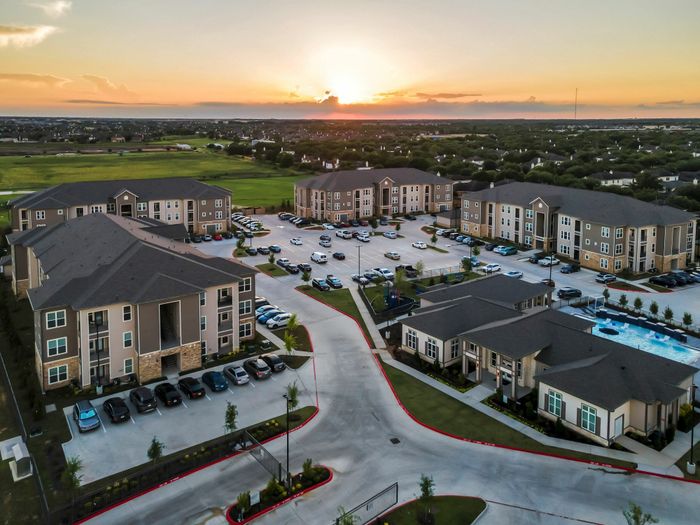 Picturesque aerial view of a modern apartment complex at sunset, featuring elegant buildings, ample parking, and a refreshing pool area enveloped by lush greenery.