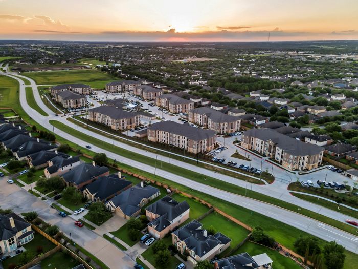 Aerial view showcasing a modern apartment complex surrounded by lush greenery and suburban homes, beautifully illuminated by a glowing sunset, highlighting community amenities and convenient road access.