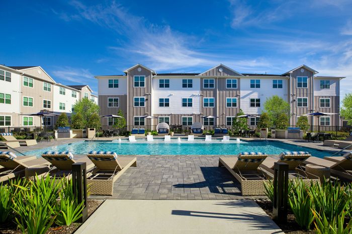 Luxurious apartment complex with a sparkling pool, stylish lounge chairs, and lush landscaping under a clear blue sky.