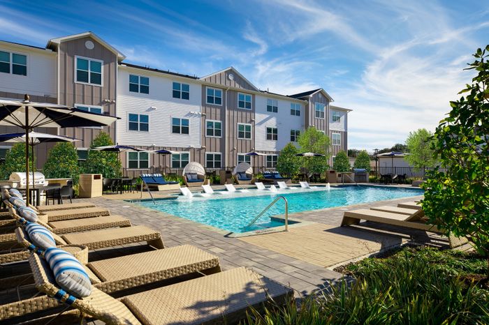 Luxurious apartment complex with a sparkling outdoor pool, stylish lounge chairs, and vibrant greenery under a bright blue sky.