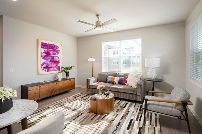 Modern living room with a sleek ceiling fan, chic furniture, vibrant artwork, large windows, and cozy seating.
