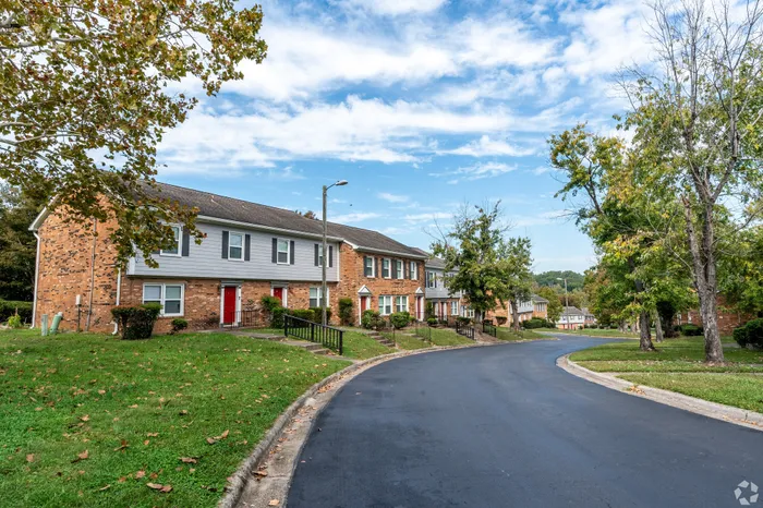 Charming suburban street with row of brick townhouses, lush green lawns, and mature trees under a bright blue sky.