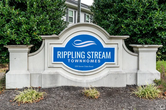 Inviting entrance sign for Rippling Stream Townhomes, set amidst lush green landscaping, showcasing elegant townhome living at 200 Seven Oaks Road.