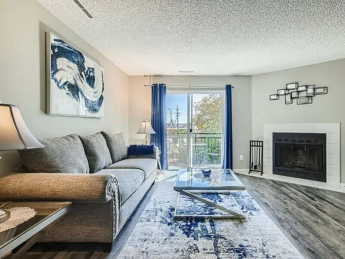 Bright and inviting living room with a modern sofa, abstract art, and a cozy fireplace, complemented by elegant blue accents and a view of the balcony.