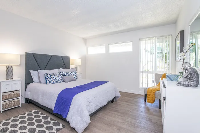 Bright and spacious bedroom featuring modern decor, a comfortable bed with stylish pillows, sleek nightstands with lamps, and a cozy seating area. Large windows allow abundant natural light, creating an inviting atmosphere.
