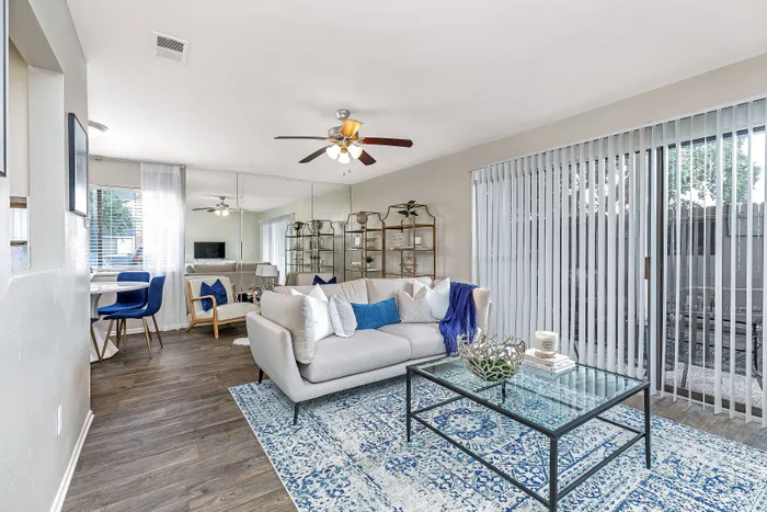 Bright and modern living room with stylish decor, featuring a cozy sofa, elegant glass coffee table, and large windows with vertical blinds offering natural light.