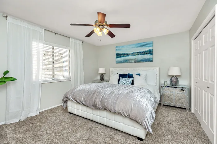 Inviting bedroom with plush carpeting, elegant bedding, and soothing natural light, featuring a ceiling fan and artwork for a serene ambiance.