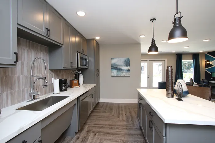 Modern kitchen with sleek gray cabinetry, elegant marble countertops, and industrial pendant lighting, leading to an inviting living area.