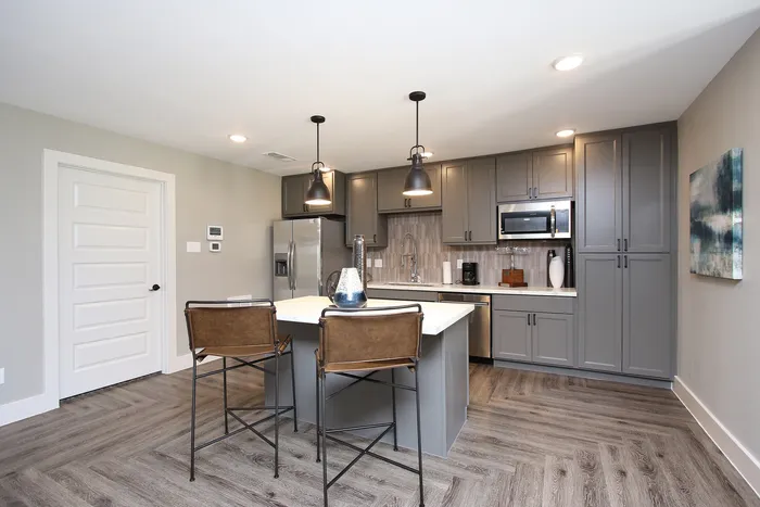 Modern kitchen with sleek grey cabinets, stainless steel appliances, and elegant pendant lighting over a central island with leather bar stools.