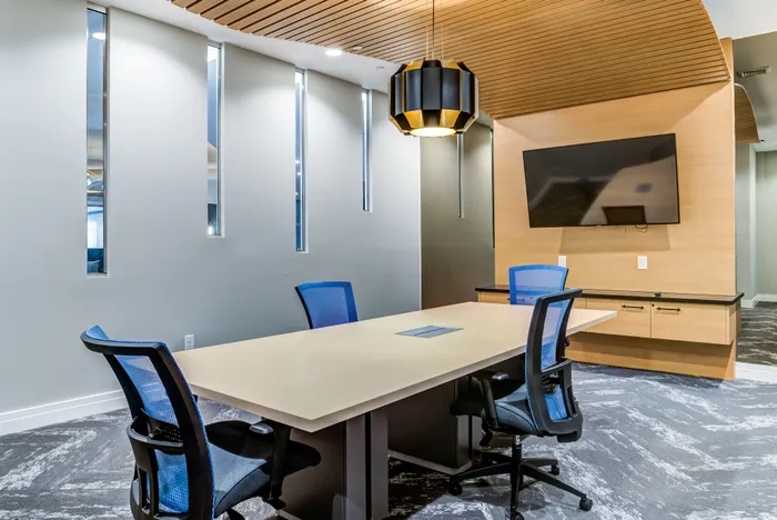 Modern conference room featuring a sleek table, vibrant blue chairs, stylish pendant lighting, and a wall-mounted TV, perfect for professional meetings.