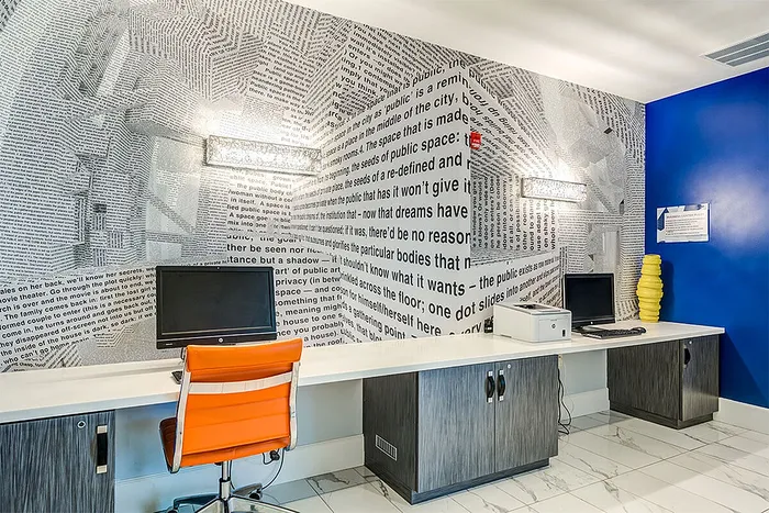 Chic workspace in modern apartment complex featuring text-inspired wall art, sleek computers, and vibrant orange chair.