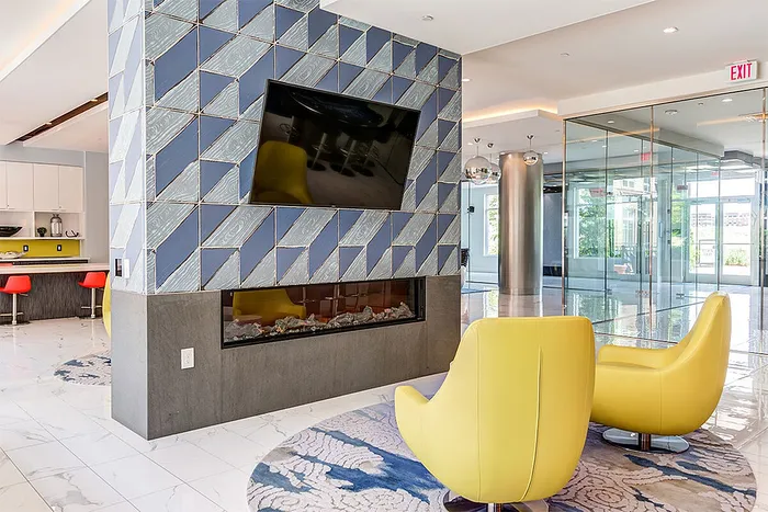 Modern apartment lobby with stylish geometric wall tiles, cozy yellow chairs, and a sleek fireplace beneath a mounted TV.