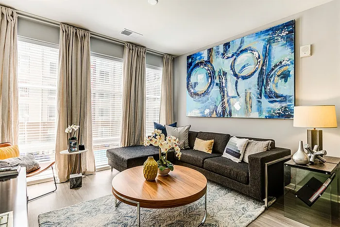 Modern living room with chic gray sofa adorned with throw pillows, a stylish wooden coffee table, elegant decorative plants, and a large abstract painting, complemented by natural light filtering through floor-to-ceiling windows.