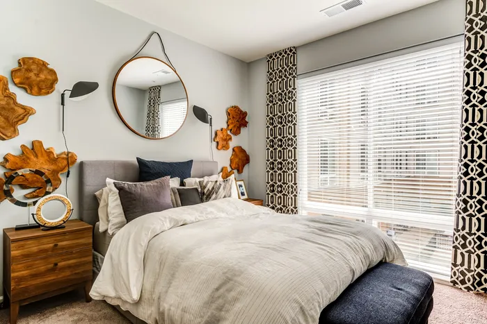 Inviting bedroom with plush bedding, elegant wood accents, a large circular mirror, and abundant natural light through stylish blinds.