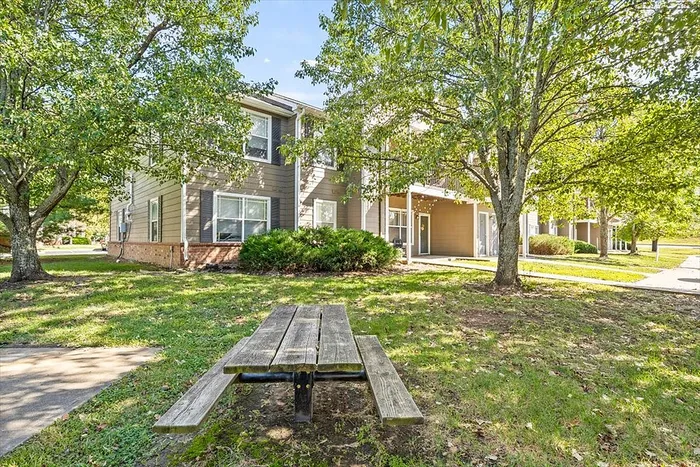 Charming two-story apartment building surrounded by lush greenery, featuring a welcoming picnic area and shaded pathways.