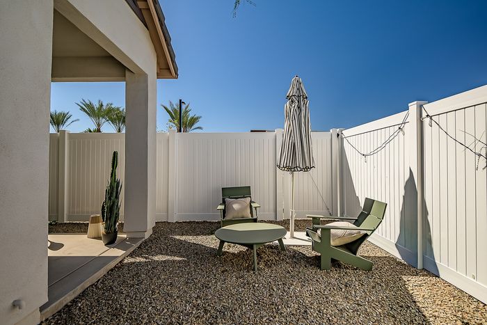 Chic outdoor patio featuring modern chairs, a stylish table, and a striped umbrella against a sunny blue sky, surrounded by a privacy fence with lush palm tree views.