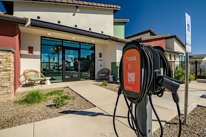 Modern apartment entrance with sleek design and electric vehicle charging station, surrounded by manicured landscape under clear blue skies.