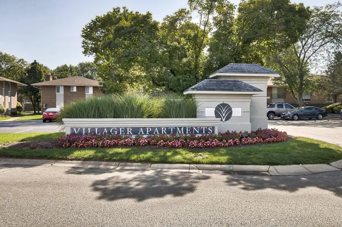 Inviting entrance to Villager Apartments, surrounded by lush greenery and vibrant floral displays, offering a serene living environment.