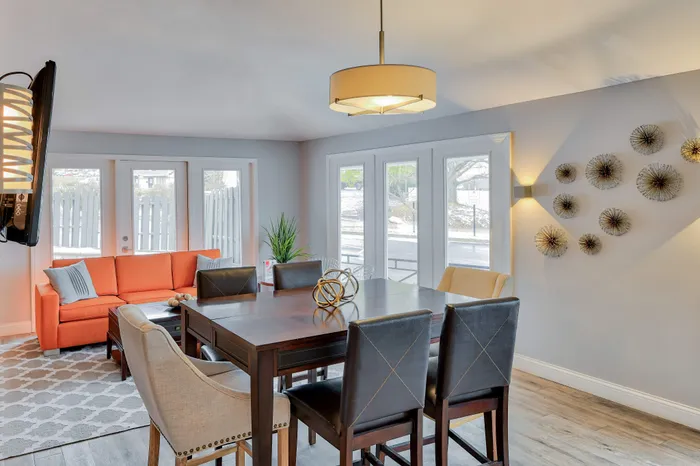 Elegant dining area with modern decor, featuring a sleek wooden table, stylish chairs, and a vibrant orange sofa by large windows with serene outdoor views.