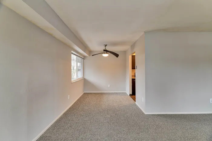 Bright and inviting apartment space featuring a textured carpet, modern ceiling fan, and ample natural light from the large window.