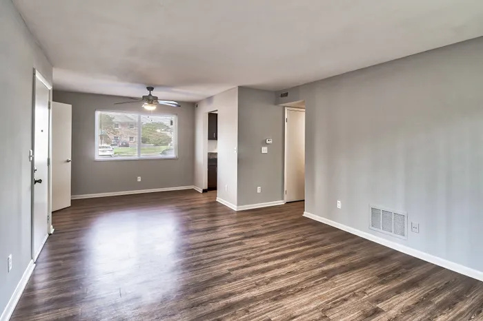 Bright open living space with modern laminate flooring, large window, ceiling fan, and neutral gray walls, perfect for versatile decorating in a cozy apartment.