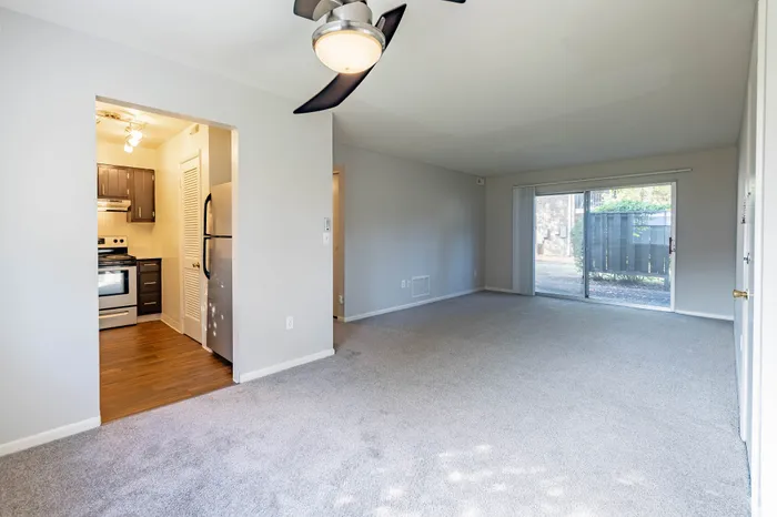 Bright and airy living space with an open connection to a sleek modern kitchen featuring stainless steel appliances and warm wood flooring, complemented by plush carpeting and a large sliding glass door leading to a serene outdoor view.