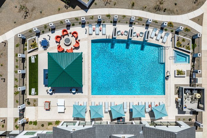 Aerial view of luxurious outdoor pool area featuring a shimmering blue swimming pool, cozy circular fire pit seating with red chairs, parasol-shaded lounge chairs, and a stylish green-roofed pavilion ideal for relaxation and entertaining.