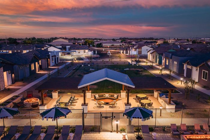 Charming neighborhood at twilight showcasing cozy homes, a beautifully lit communal area with tables and chairs, and a warm sunset sky backdrop.