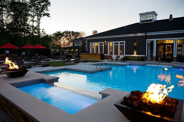 Luxurious apartment complex featuring a stunning pool area with ambient fire bowls, surrounded by stylish lounge chairs and vibrant red umbrellas at sunset.
