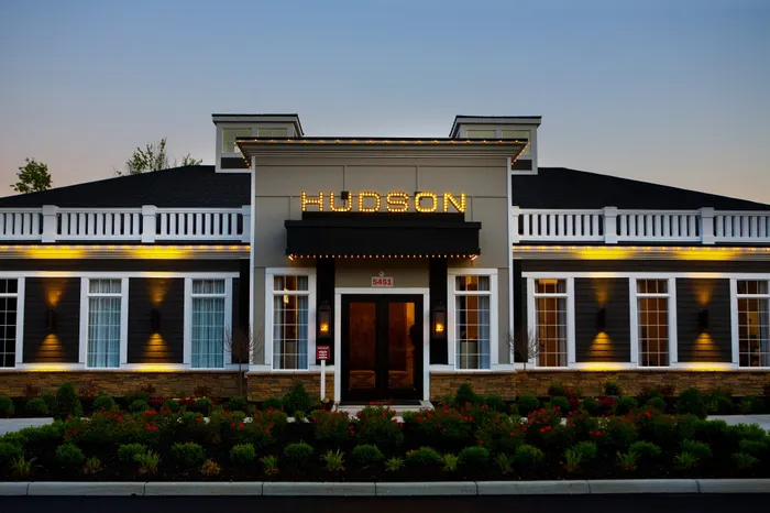 Elegant and modern building facade at dusk, featuring 'Hudson' sign illuminated with vibrant lights, surrounded by lush landscaping.