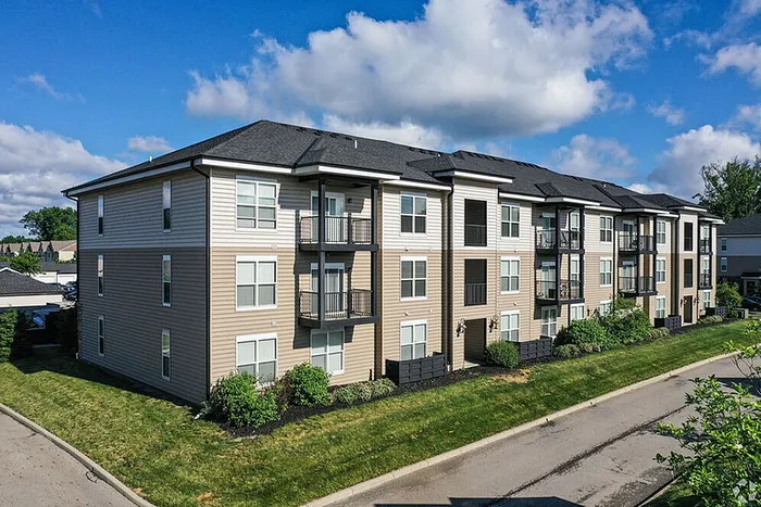 Modern three-story apartment building with balconies, surrounded by lush greenery under a bright blue sky.
