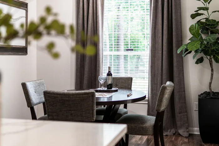 Chic dining area with elegant round table, wine setup, and lush greenery by a sunlit window.