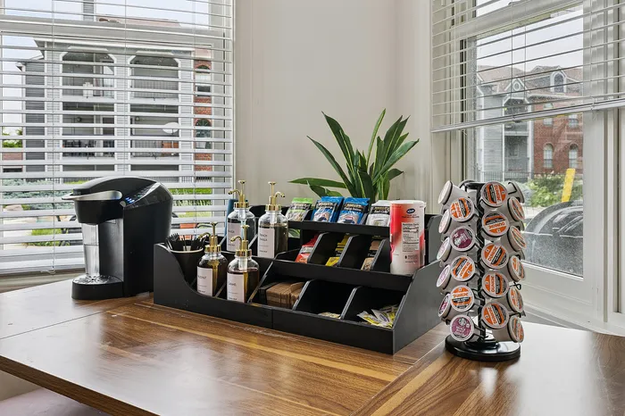 Inviting coffee nook with modern Keurig setup, flavor syrups, and assorted pods, framed by bright windows and a leafy green plant.