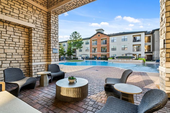 Luxurious apartment complex featuring a sparkling pool view from a shaded outdoor seating area, surrounded by beautiful architecture and lush greenery.