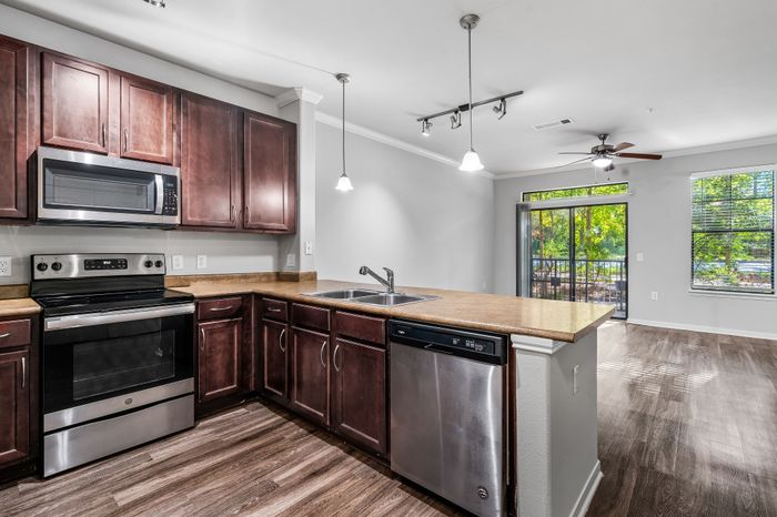Modern kitchen with rich wood cabinetry, stainless steel appliances, and a luminous open-concept living area with large windows overlooking lush greenery.