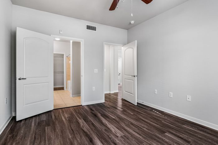 Modern apartment interior featuring sleek wood flooring, light gray walls, and a spacious open doorway leading to a bright bathroom with tiled surfaces.