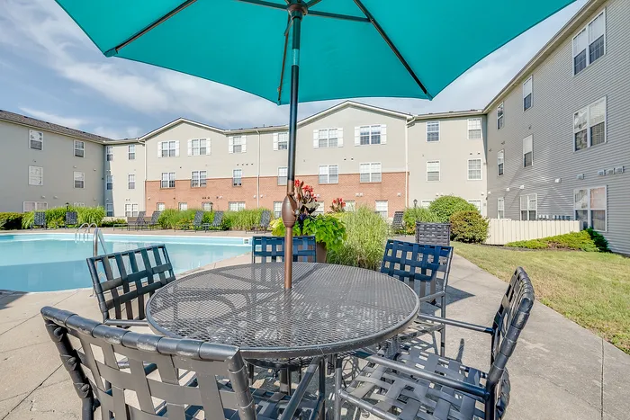 Inviting outdoor swimming pool area with elegant patio seating under a vibrant blue umbrella, surrounded by lush greenery and modern apartment buildings.