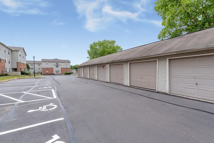 Scenic view of a modern apartment complex with a row of spacious garages, ample parking, and lush greenery under a clear blue sky.