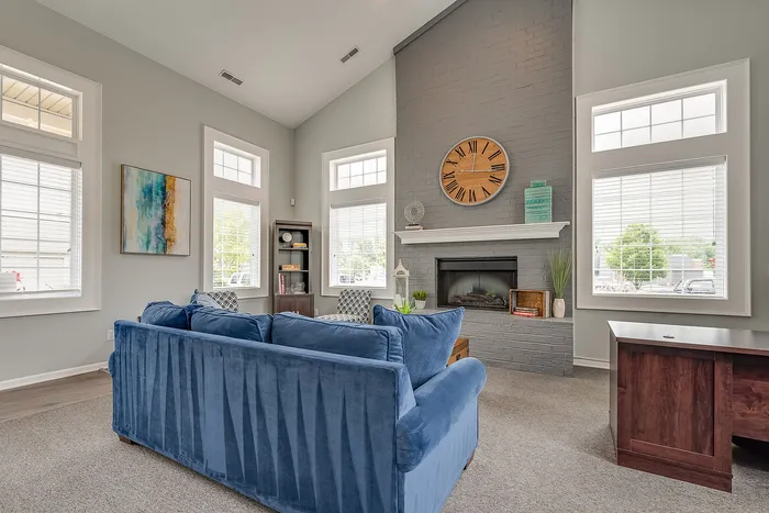 Bright and inviting living room with large windows, a cozy blue sofa, and a stylish brick fireplace adorned with a rustic clock.