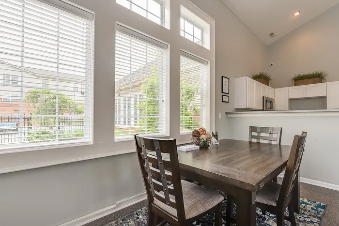 Bright dining area with modern wooden furniture and large windows offering scenic views, adjacent to a sleek kitchen with white cabinetry.