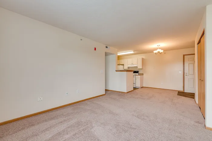 Bright and open living space with plush carpet, adjacent to a modern kitchen with white cabinetry and ample natural lighting.