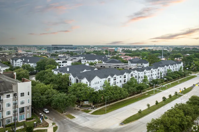 Elevated view of a modern apartment complex surrounded by lush greenery and wide streets under a serene sky.