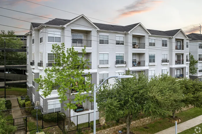 Modern three-story apartment complex with private balconies, surrounded by lush greenery, set against a picturesque evening sky.