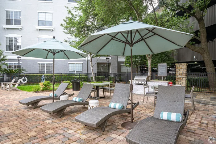 Inviting outdoor lounge area featuring stylish wicker chaise lounges and shaded umbrellas, set amidst lush greenery and a modern barbecue grill station.