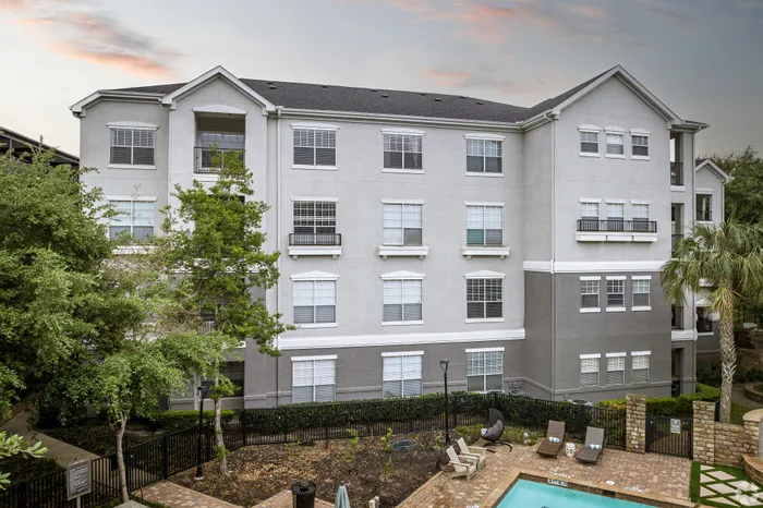 Elegant apartment building exterior with large windows, surrounded by lush greenery, featuring a charming patio area with lounge chairs and a pool.