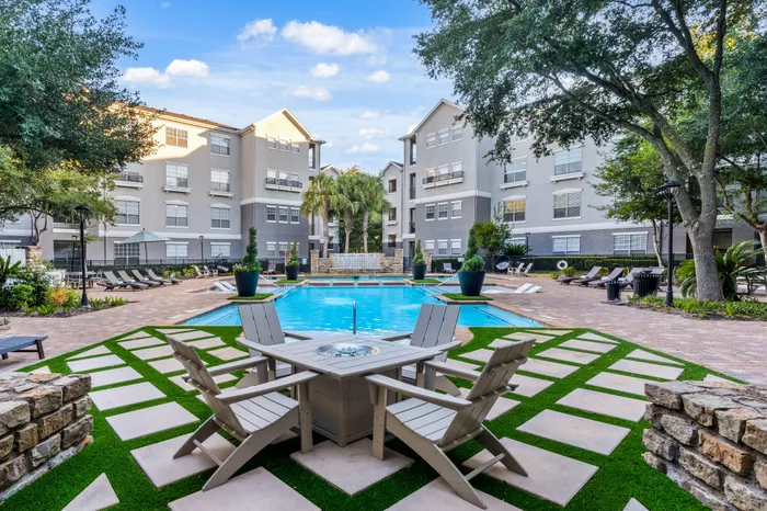 Luxurious apartment complex courtyard featuring a sparkling pool, elegant patio seating, and lush landscaping under a vibrant blue sky.