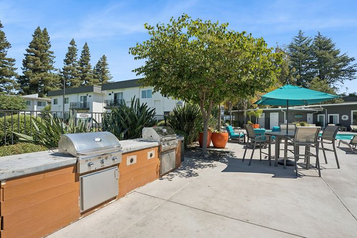 Charming outdoor common area featuring built-in stainless steel BBQs, stylish patio seating with teal umbrellas, and lush greenery, perfect for social gatherings.